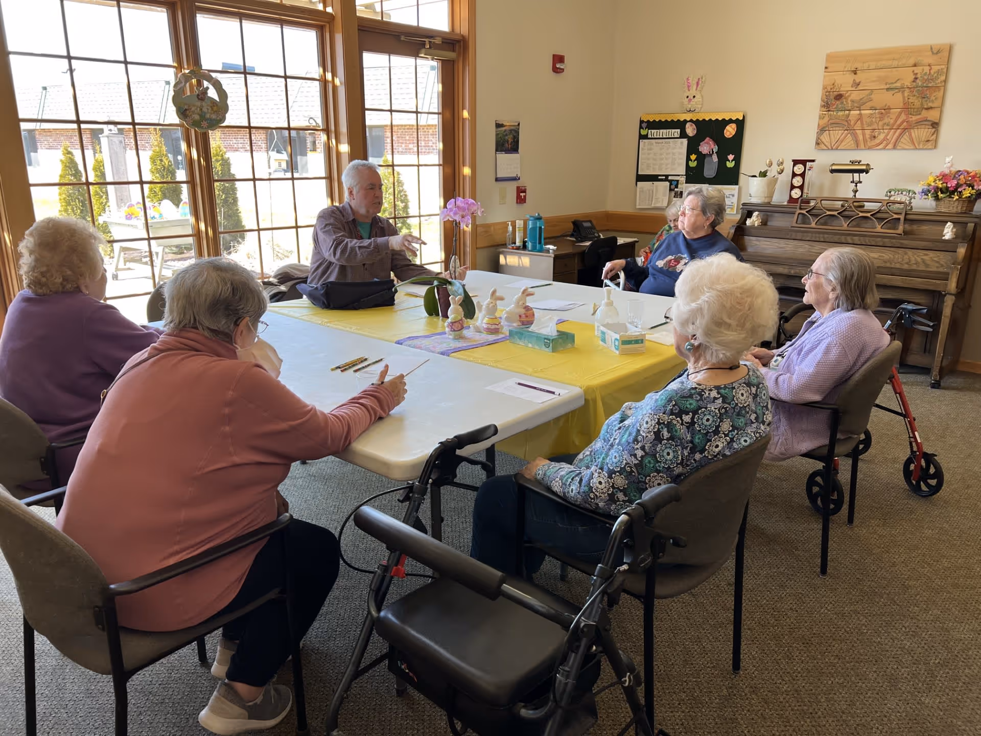 A group of elderly people sitting around a table in a well-lit room with large windows. The table is covered with a yellow tablecloth and has Easter-themed decorations, tissues, and hand sanitizer on it. There is a piano against the wall and a bulletin board with activities listed. The room appears to be a communal space for activities or meetings.