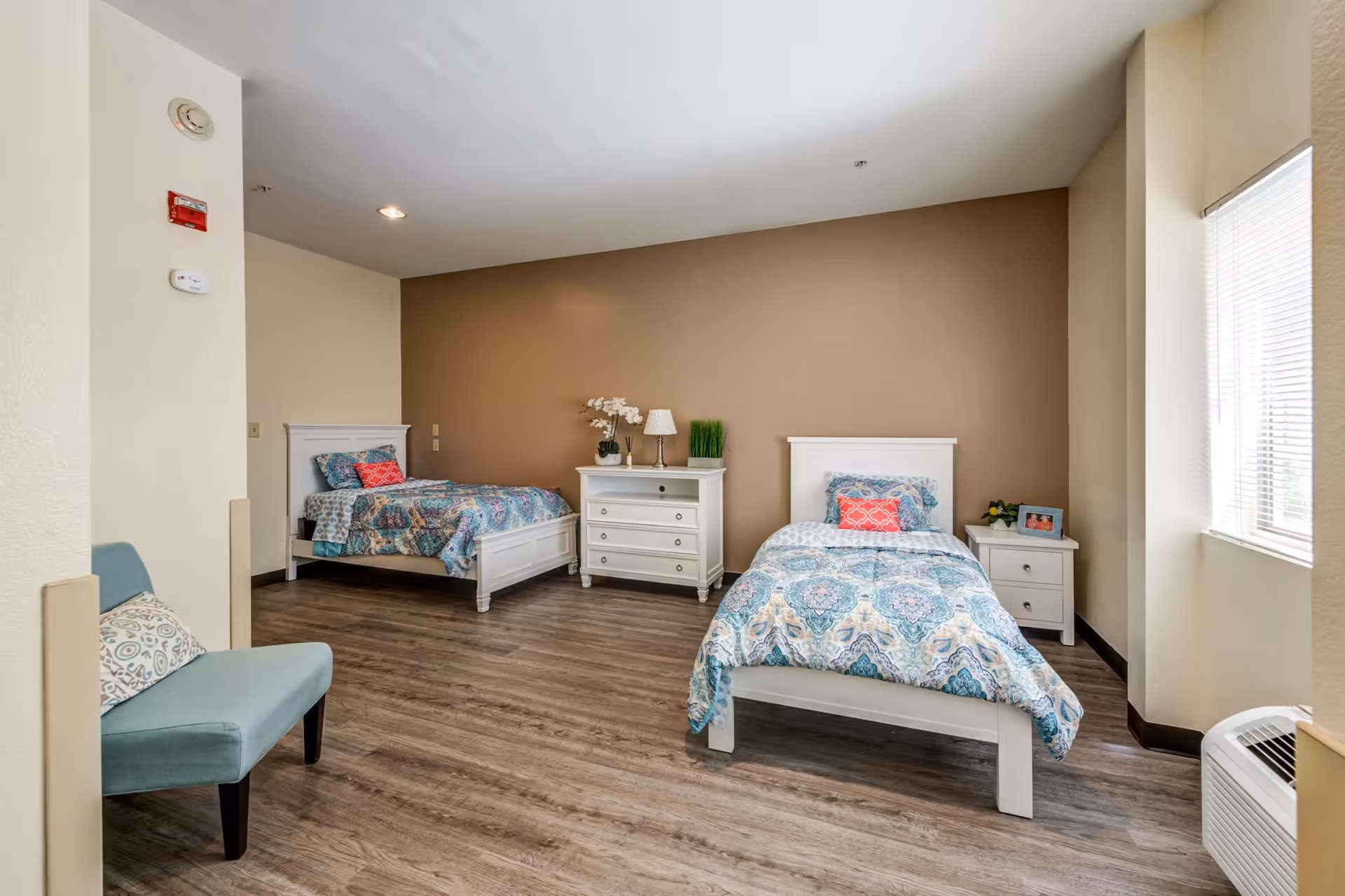 A bright and clean bedroom with two single beds, each with patterned blue and white bedding and a red accent pillow. Between the beds is a white dresser with decorative items including a lamp, a plant, and flowers. There is a small white nightstand with a framed photo and a plant next to the bed on the right. A light blue chair with a patterned pillow is positioned near the left side of the room. The room has wood flooring, beige walls, and a large window with blinds letting in natural light.