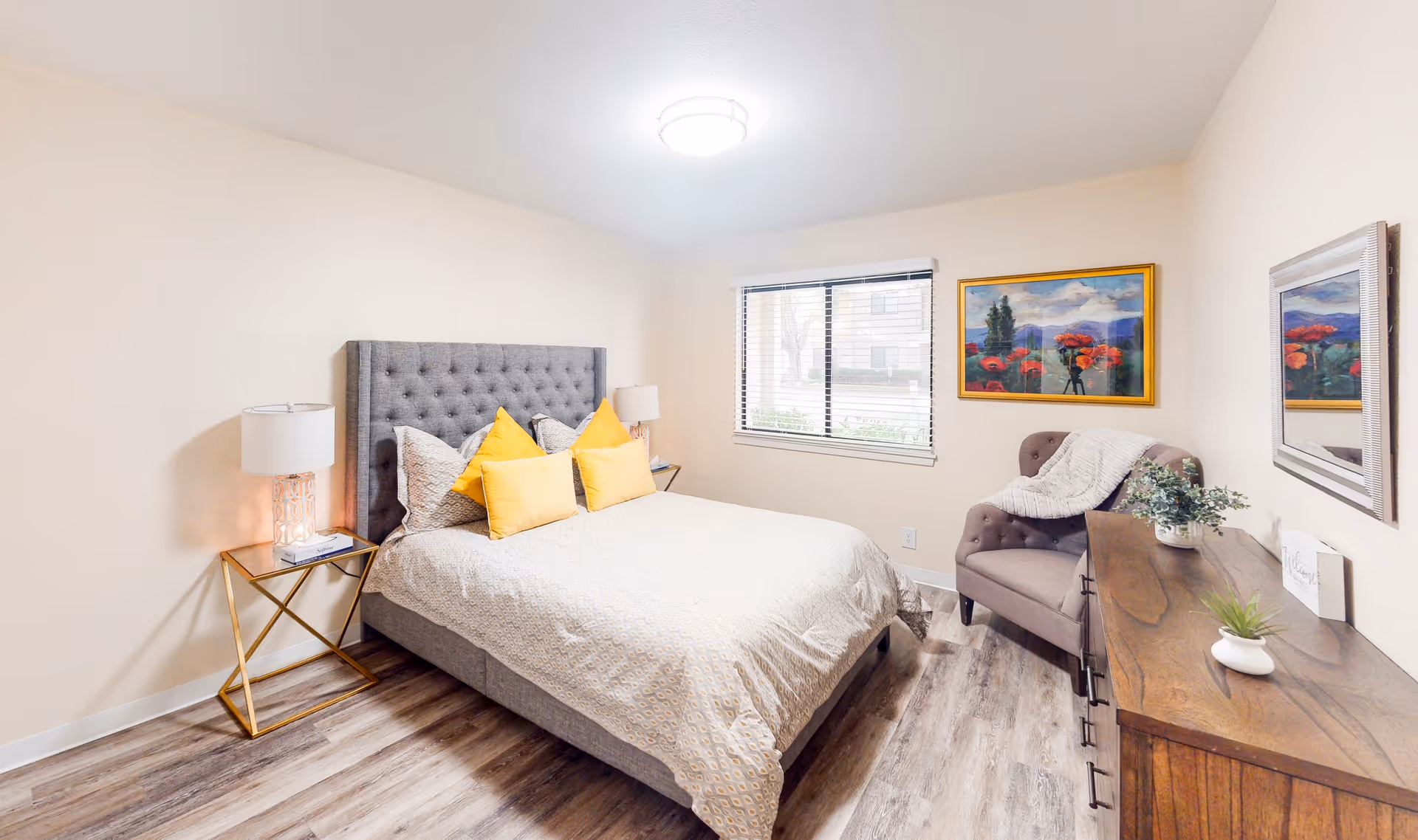 A bright and cozy bedroom with a large gray upholstered bed adorned with yellow and gray pillows. There is a wooden nightstand with a lamp on the left side of the bed. A window with blinds lets in natural light. On the right side of the room, there is a cushioned armchair with a throw blanket, a wooden dresser with decorative plants, a mirror above it, and a colorful framed painting on the wall.