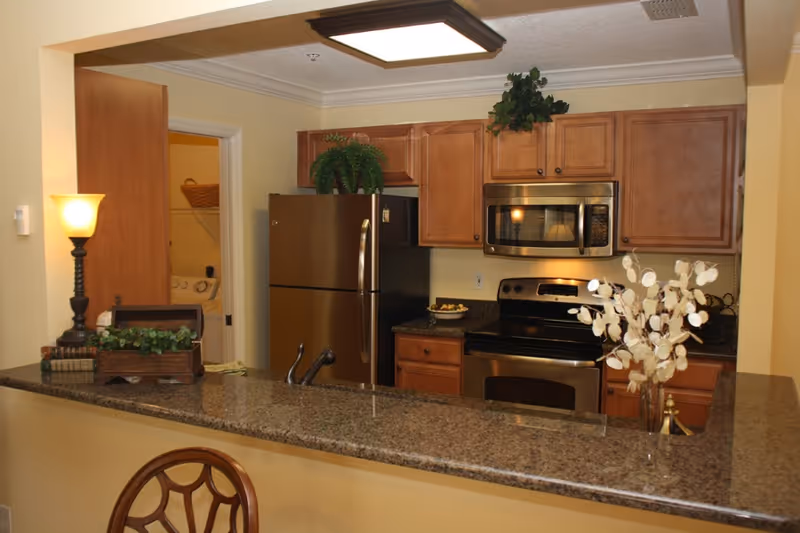 A kitchen area with wooden cabinets, a stainless steel refrigerator, microwave, and stove. The countertop is granite with a sink and decorative items including a lamp, a small plant, and a vase with white flowers. An open door reveals a laundry room with a washer and dryer.