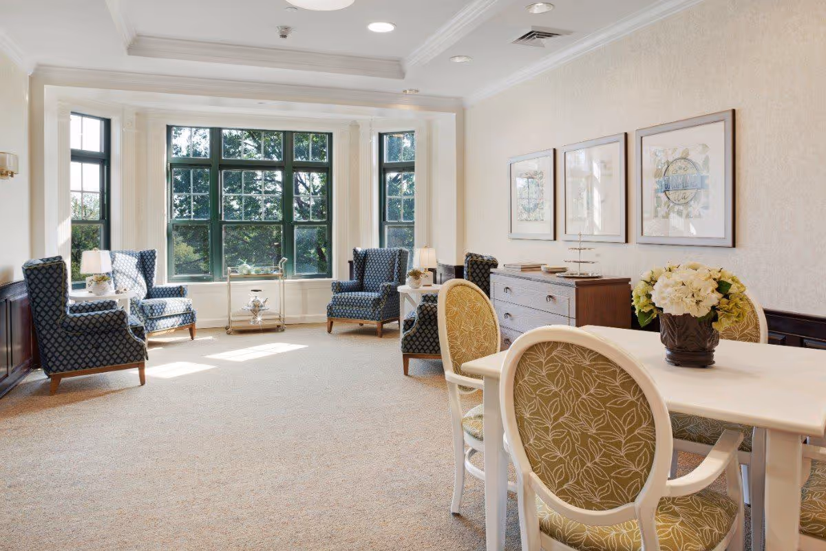 A bright and elegant sitting room with large windows letting in natural light. The room features four blue patterned armchairs arranged near the windows, a white table with four green patterned chairs, a wooden sideboard with framed artwork above it, and a vase of white flowers on the table.