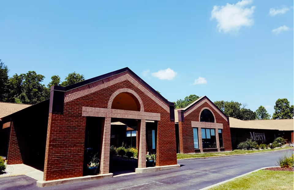 Front exterior of a red-brick Mercy Rehab and Care Center with a covered entrance, driveway, and clear blue sky.