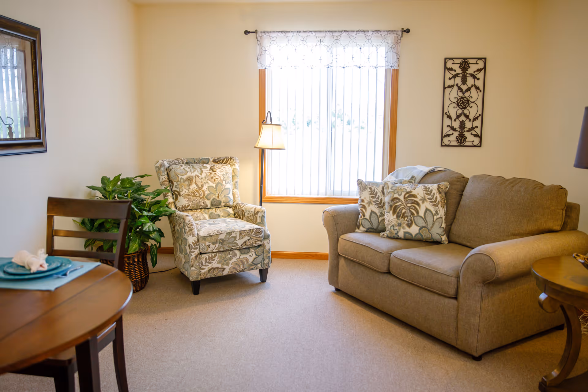 A cozy living room area with a beige couch adorned with patterned cushions, a floral upholstered armchair, a floor lamp, a wooden side table, a round wooden dining table with a chair, and a potted plant near the corner. A window with vertical blinds and a decorative valance lets in natural light, and a framed mirror and wall art decorate the walls.