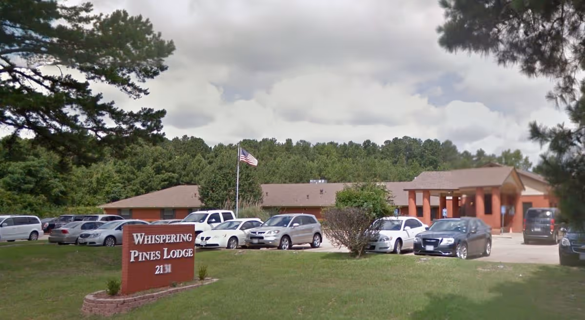 Exterior view of Whispering Pines Lodge, a single-story building with a brown roof surrounded by trees. Several cars are parked in front of the building, and an American flag is flying on a flagpole near the entrance. A red sign with white lettering displays the facility's name and address.