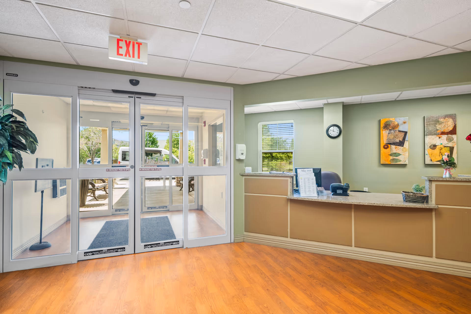 Reception area of Serenity Estates of Morris featuring a front desk with a computer, phone, and decorative items. Behind the desk is a green wall with two abstract paintings and a clock. To the left are glass automatic doors leading outside, with an exit sign above. The floor is wooden and there is a potted plant near the entrance.