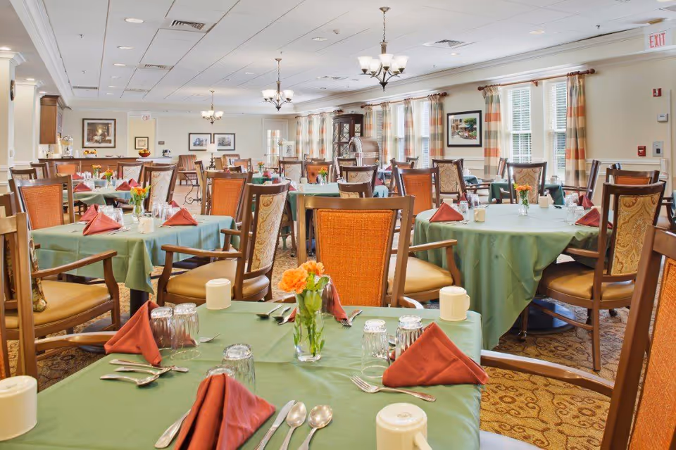 A bright and spacious dining room with multiple tables covered in green tablecloths, each set with red folded napkins, glasses, silverware, and small flower arrangements. The room has large windows with striped curtains, framed artwork on the walls, and elegant chandeliers hanging from the ceiling.