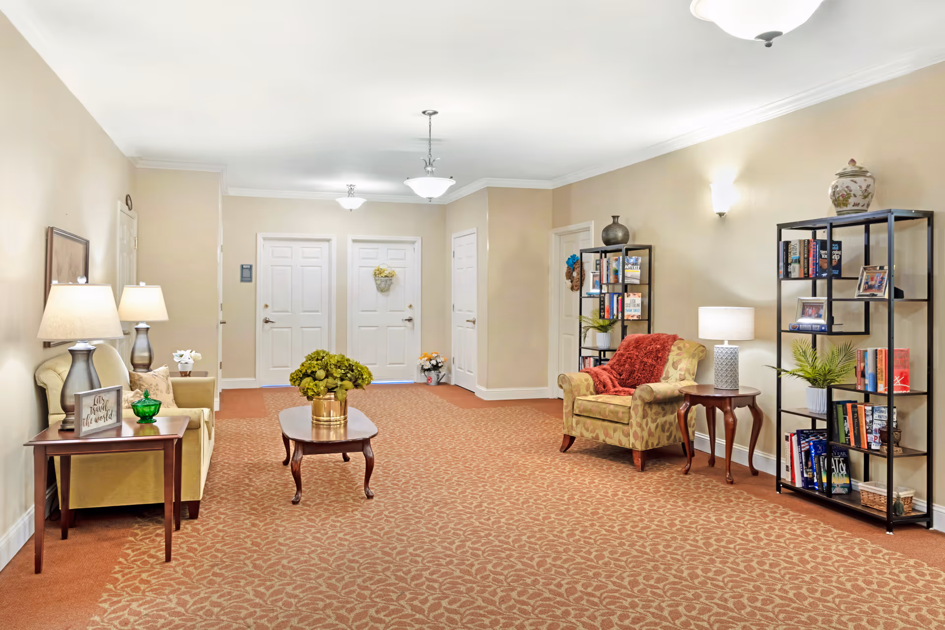 Well-lit senior living common lounge with armchairs, a coffee table, bookshelves, and doors down a hallway.