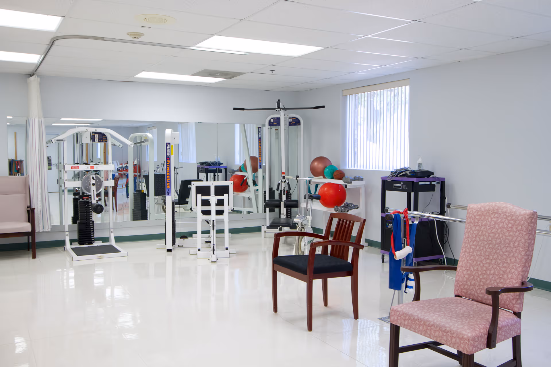 A bright exercise room in a senior living facility with various fitness equipment including weight machines, exercise balls, and dumbbells. The room has large mirrors on one wall, two chairs in the foreground, and a window with vertical blinds letting in natural light.