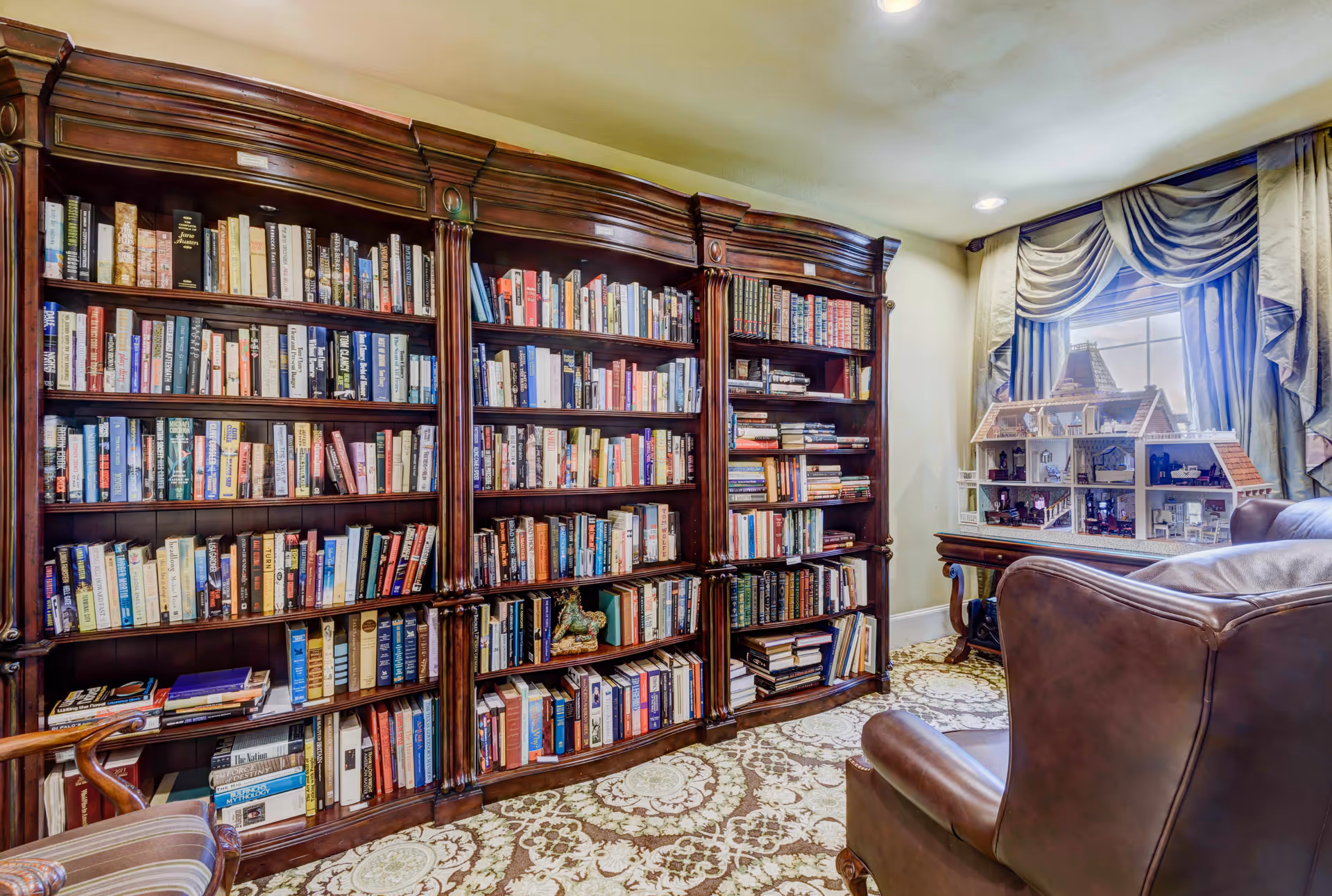 A cozy library room with a large wooden bookshelf filled with books, a patterned carpet, a leather armchair, and a table holding a detailed dollhouse near a window with draped curtains.