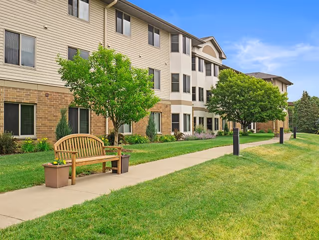 Exterior view of a multi-story senior living facility building with beige siding and brick accents. There is a concrete walkway in front of the building, flanked by green grass, trees, and a wooden bench with flower pots on either side. The sky is clear and blue.