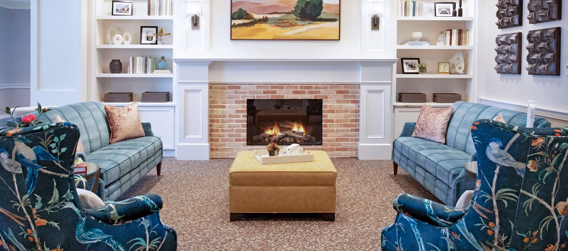 A cozy living room with two teal sofas and two armchairs with bird patterns facing a brick fireplace with a fire burning. There is a yellow ottoman in the center with a tray on top. Built-in white shelves flank the fireplace, decorated with books, framed photos, and decorative objects. A landscape painting hangs above the fireplace.