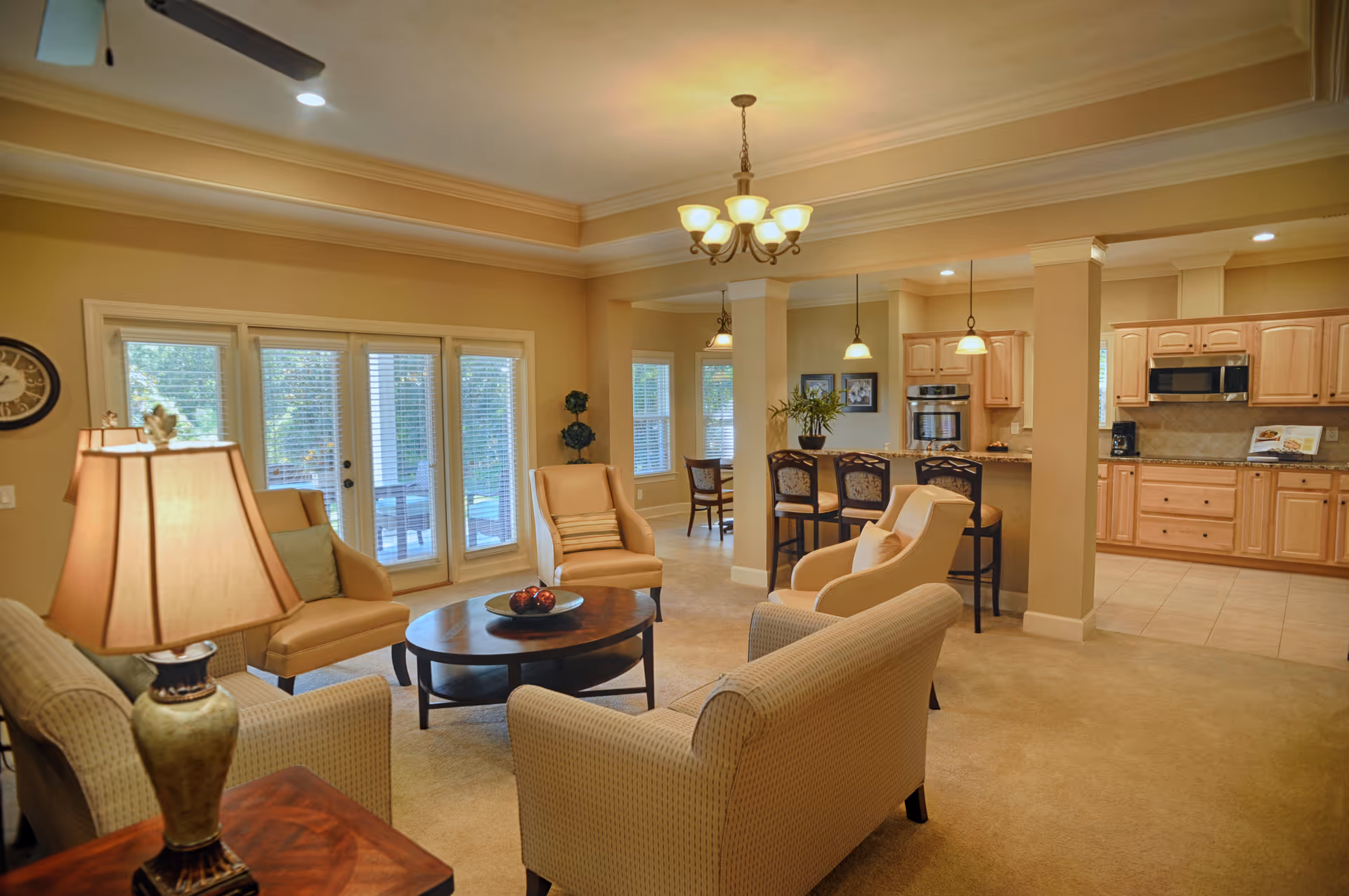 Open-plan living room with beige sofas and armchairs around a round coffee table, leading to a kitchen with a breakfast bar and pendant lights.