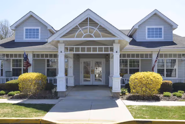 Front entrance of a gray building with a covered portico, white columns, double glass doors, dormer windows, American flags, and landscaped shrubs.