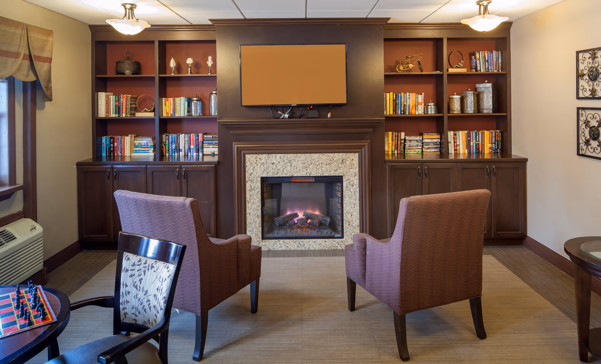 Cozy common room with two armchairs facing a fireplace flanked by built-in bookshelves and a wall-mounted TV.