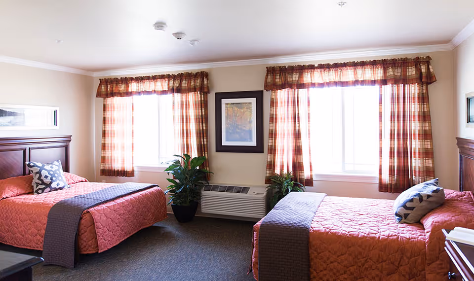 A bright bedroom with two beds covered in red quilted bedspreads and gray throws. Each bed has a decorative pillow. The room features two large windows with plaid curtains, a framed picture on the wall between the windows, and two potted plants near a wall-mounted air conditioning unit.