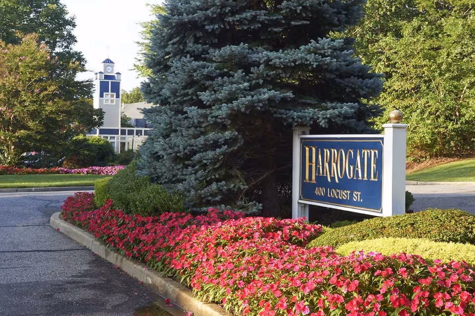 Entrance to Harrogate Retirement Community featuring a landscaped area with vibrant pink flowers, green bushes, a large evergreen tree, and a blue and white sign displaying 'HARROGATE 400 LOCUST ST.' A building with a clock tower is visible in the background.