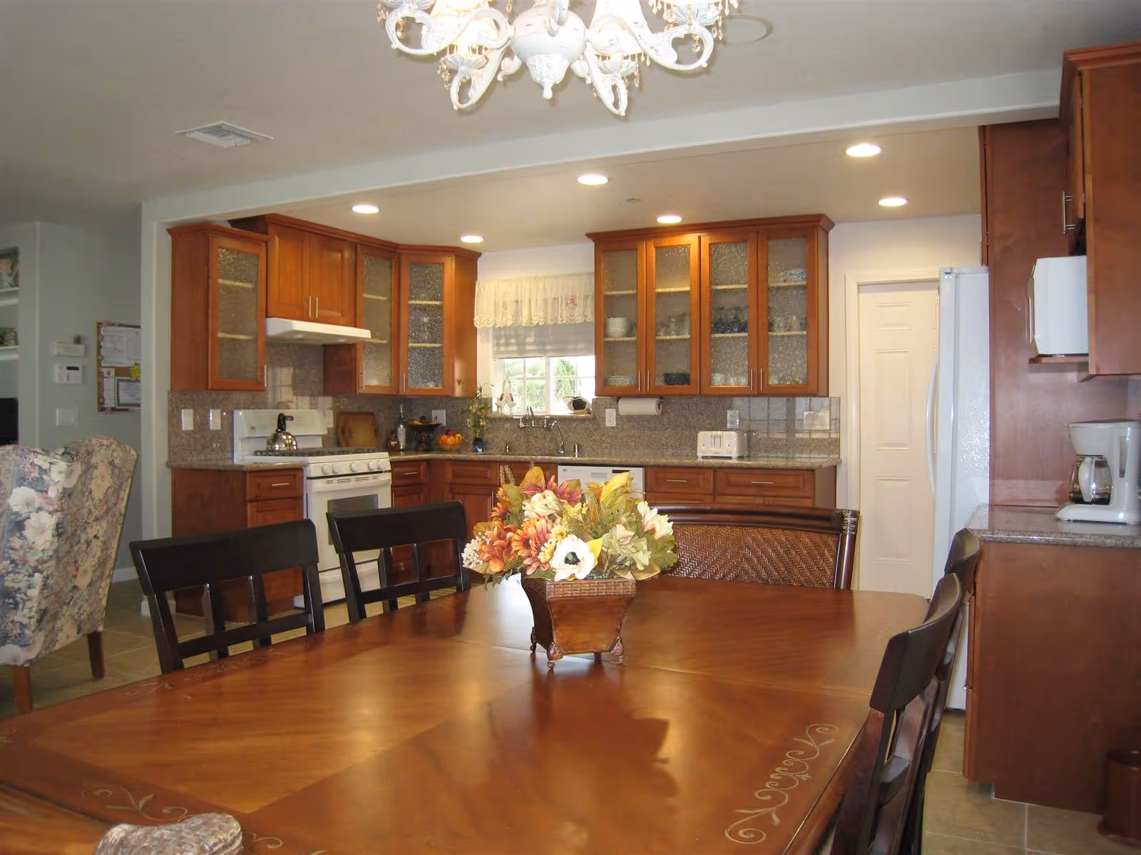 A well-lit kitchen with wooden cabinets, a white stove, a refrigerator, a microwave, and a coffee maker. In the foreground, there is a wooden dining table with a floral centerpiece and several chairs around it. A floral upholstered chair is partially visible to the left.