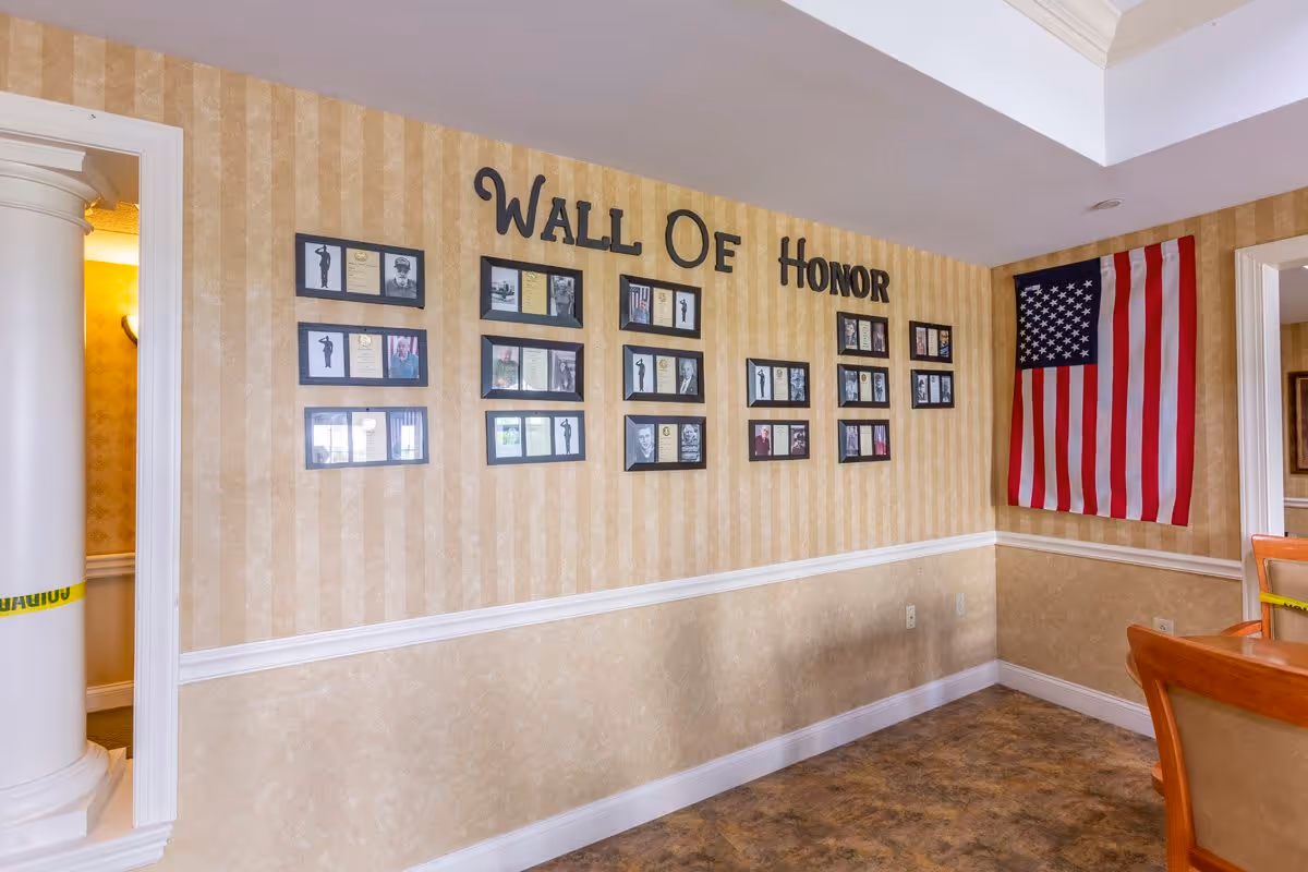 Interior wall of a senior living facility with a 'Wall of Honor' display featuring framed photos and plaques. An American flag is hanging on the right side of the wall. The room has beige striped wallpaper with a white chair rail and a brown floor. Part of a wooden chair is visible on the right side.