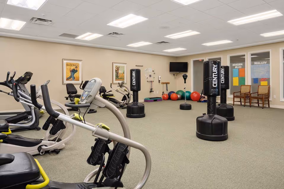 A spacious fitness room with exercise equipment including stationary bikes and elliptical machines. The room also features three black Century punching bags, colorful exercise balls, and various fitness accessories. The walls are decorated with framed vintage-style posters and there are windows with signs reading 'MILEAGE CLUB'. Two wooden chairs are placed near the windows.