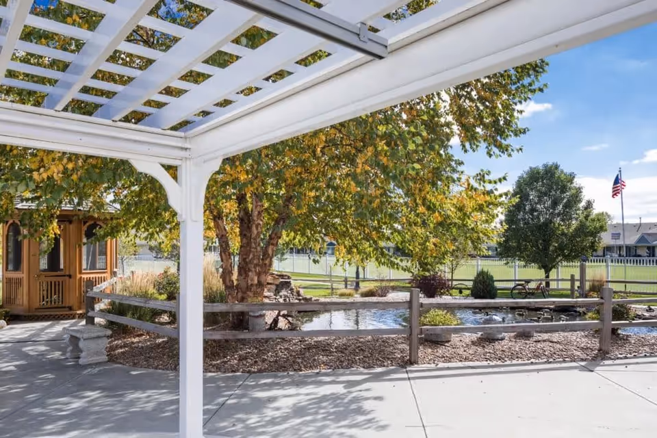 View from under a white pergola overlooking a landscaped garden area with a small pond, trees with green and yellow leaves, a wooden gazebo, and an American flag in the background on a sunny day.
