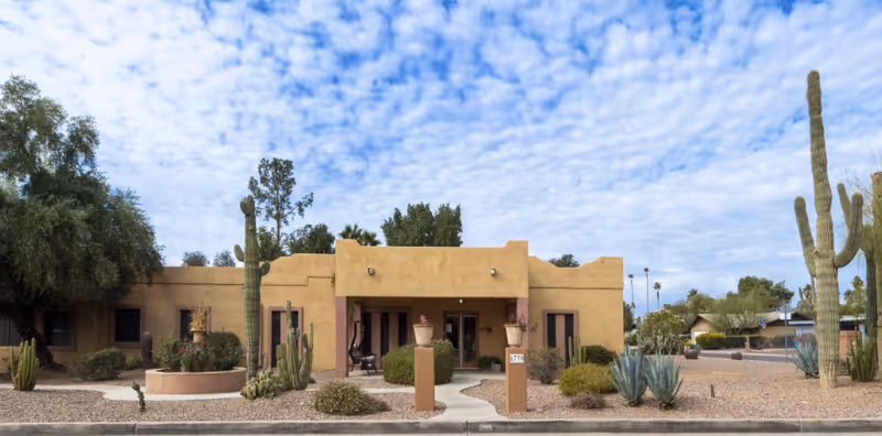 Exterior front view of a single-story adobe-style building with desert landscaping including cacti and shrubs under a partly cloudy sky.