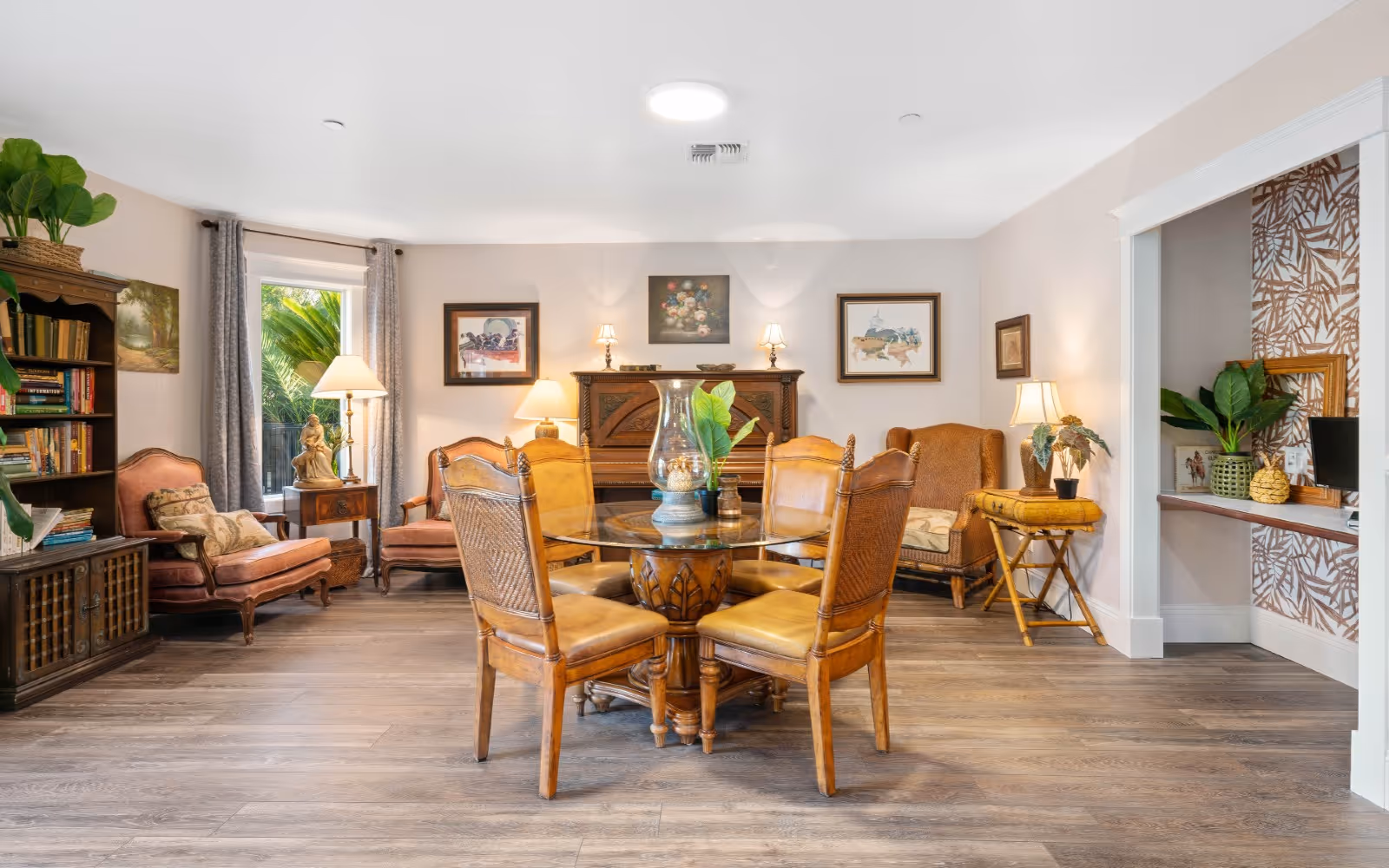 A cozy living room with wooden flooring featuring a round glass-top table surrounded by four wooden chairs with leather seats. The room has several armchairs and a loveseat upholstered in warm tones, a wooden bookshelf filled with books, and various lamps providing soft lighting. There are framed paintings on the walls and a small desk area with tropical-patterned wallpaper in an alcove to the right. Green plants add a touch of nature to the space.