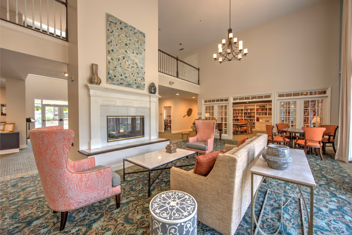A spacious and well-lit living room area in a senior living facility featuring a fireplace with a decorative painting above it, two coral patterned armchairs, a beige sofa with orange pillows, a marble coffee table, and a side table with a decorative vase. In the background, there is a room with bookshelves and tables visible through glass-paneled doors. The room has high ceilings with a chandelier and large windows with curtains.