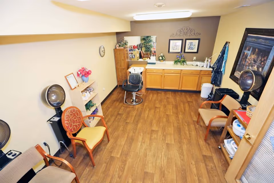 A small salon room with wooden flooring, featuring two hair drying chairs with hooded dryers, several wooden chairs with cushions, a black salon chair in front of a sink, a wooden cabinet with a large mirror above it, and decorative items including framed pictures, flowers, and plants.