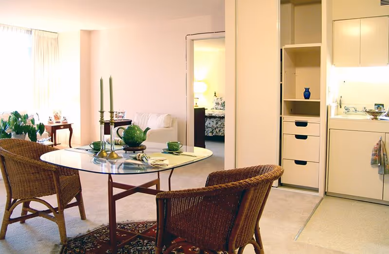 Bright senior apartment living/dining area with a glass-top table and wicker chairs, a small kitchenette to the right, and a bedroom visible through a doorway.