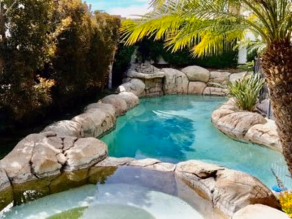 A serene outdoor pool area surrounded by large natural rocks and lush greenery including palm trees and bushes under a partly cloudy sky.