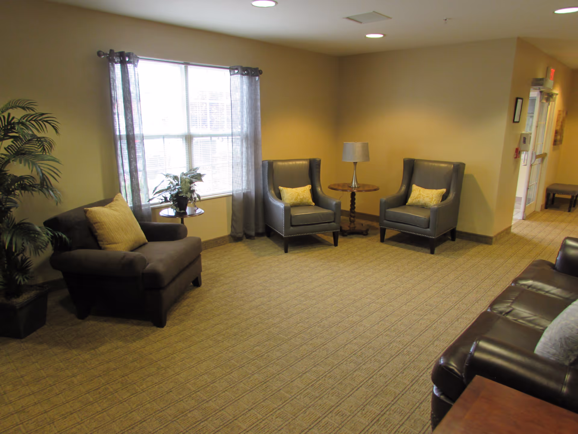 A cozy seating area in a senior living facility with two gray armchairs, a brown armchair, a brown leather sofa, a small round wooden table with a lamp, and a window with sheer curtains letting in natural light. There is also a potted plant near the window and beige carpeted flooring.