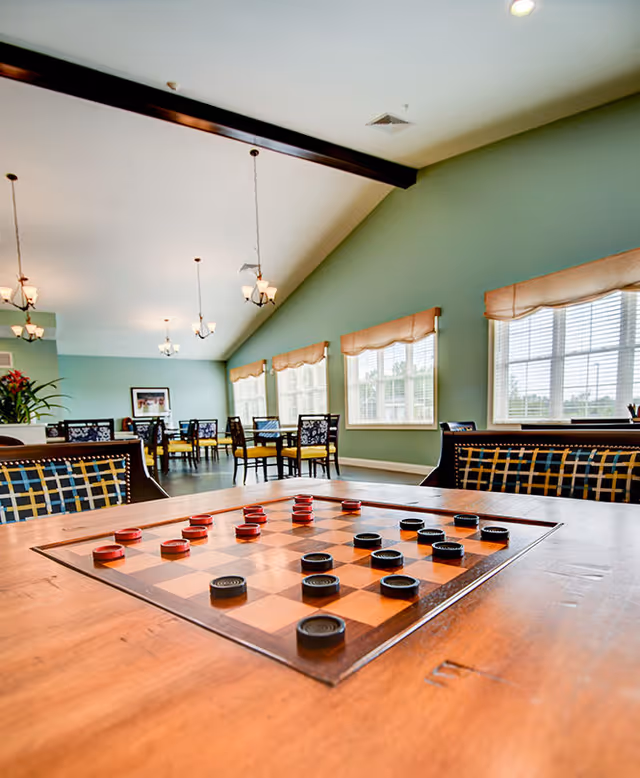 A spacious, well-lit common area with a wooden table featuring a built-in checkerboard game. The room has multiple tables and chairs with patterned upholstery, large windows with beige valances allowing natural light, and hanging light fixtures from a high ceiling with exposed beams.