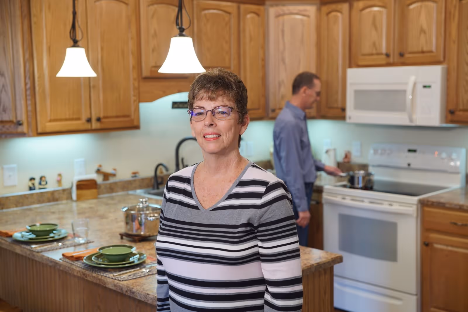 A woman wearing glasses and a striped shirt stands in a kitchen with wooden cabinets and granite countertops. In the background, a man is cooking at a stove with a pot. The kitchen is well-lit with pendant lights hanging from the ceiling and has a microwave above the stove.