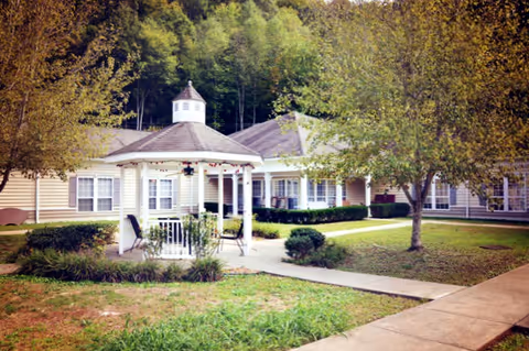 Outdoor view of Cedar Creek Assisted Living facility showing a white gazebo with chairs on a concrete path surrounded by grass, trees, and beige buildings with white trim in the background.
