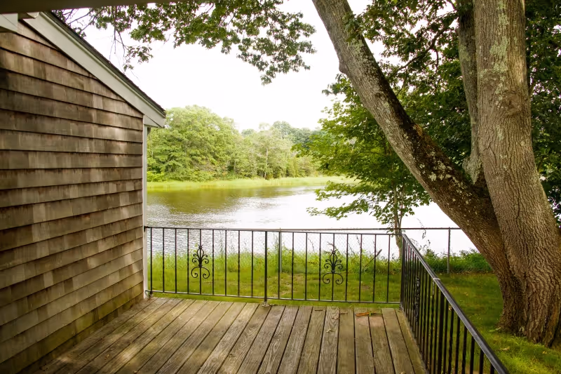 View from a wooden deck with black metal railing overlooking a calm river surrounded by green trees and grass. A large tree is visible on the right side of the deck, and the side of a wooden building is on the left.