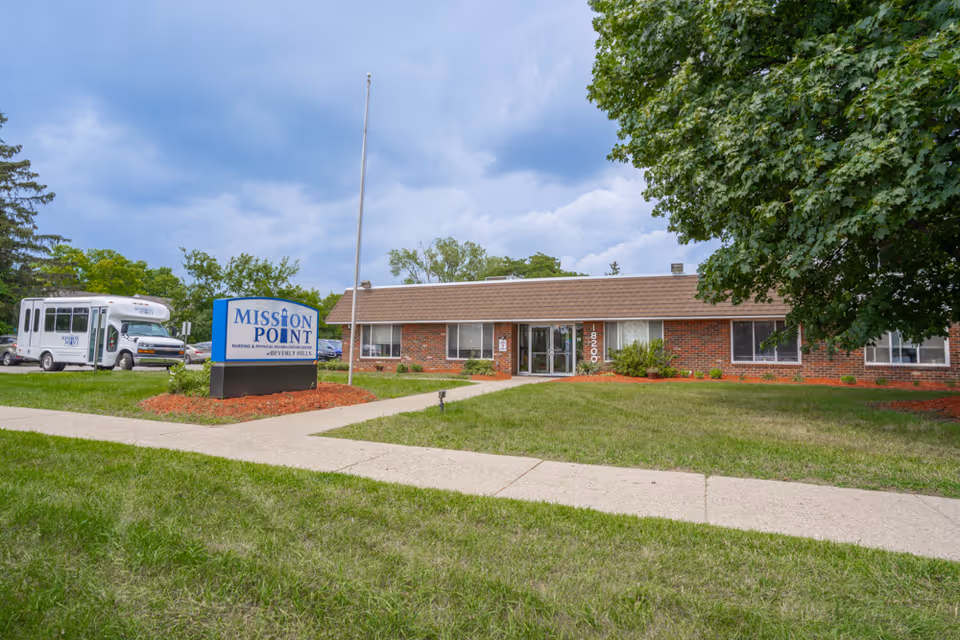 Front exterior of Mission Point Nursing & Physical Rehabilitation Center with its sign, lawn, entrance, and a parked shuttle van.