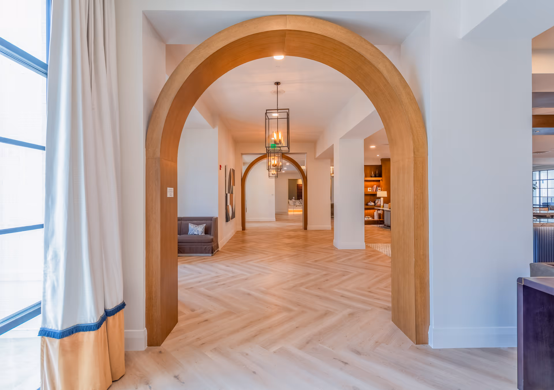 Interior view of a hallway with wooden archways and light wood herringbone flooring. There are hanging lantern-style light fixtures and white walls. A window with white curtains is visible on the left side, and a small seating area with a couch is partially visible in the background.