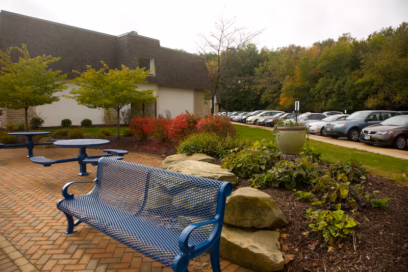 Outdoor courtyard with a blue metal bench and picnic tables, landscaped rocks and shrubs, and a row of parked cars beside a building.