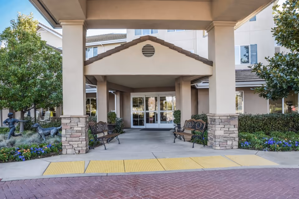 Covered entrance/portico of a senior living building with benches, landscaping, and glass double doors.