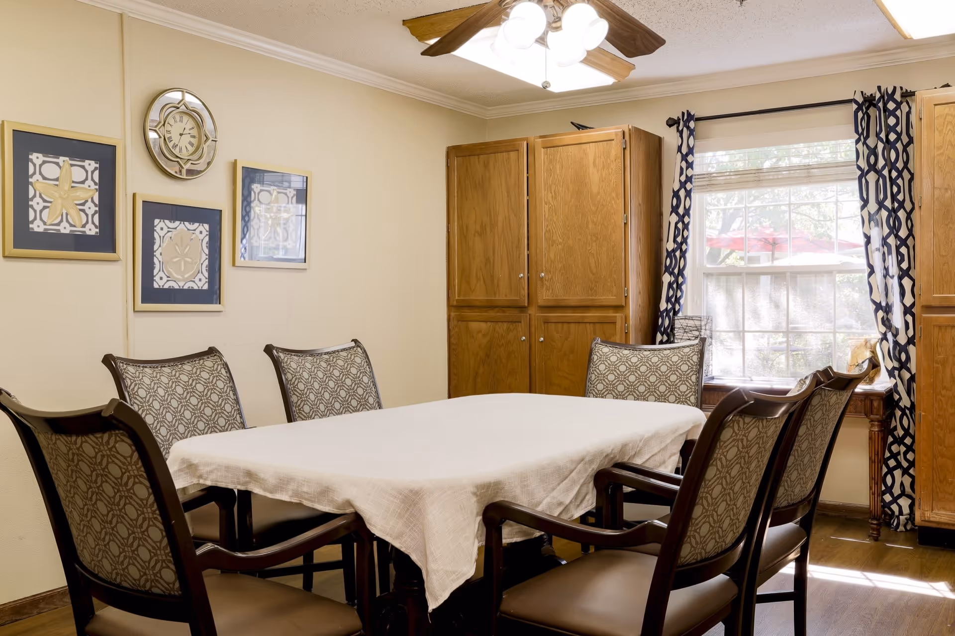 A dining room with a rectangular table covered with a white tablecloth surrounded by six upholstered chairs. The walls are decorated with three framed pictures and a round wall clock. There are wooden cabinets and a window with patterned curtains letting in natural light.