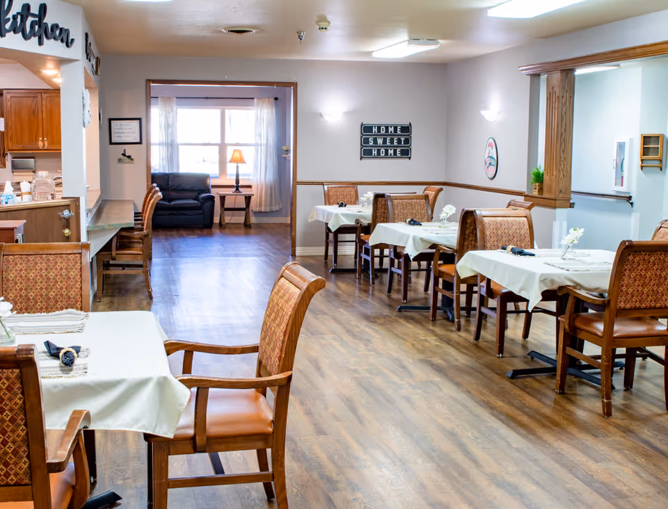 A dining room in a senior living facility with several tables covered with white tablecloths and set with napkins and small flower arrangements. Wooden chairs with patterned upholstery surround the tables. The room has wood flooring and light-colored walls with a 'Home Sweet Home' sign on one wall. In the background, there is a small seating area with a black armchair and a side table with a lamp near a window with sheer curtains. The kitchen area is partially visible on the left side.