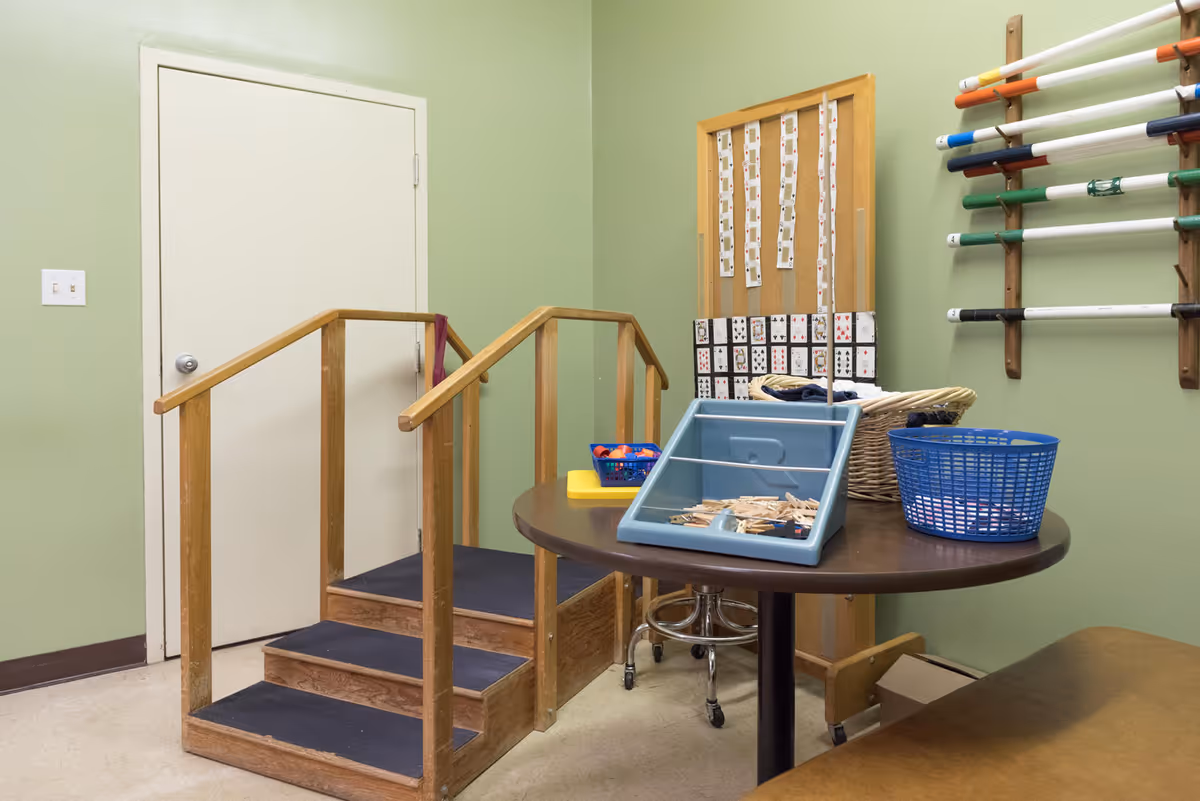 A small room with light green walls featuring a wooden three-step staircase with handrails, a round table with various activity items including a blue basket, a blue tray with clothespins, and a small basket. On the wall, there is a corkboard with playing cards pinned to it and a rack holding multiple colored rods.