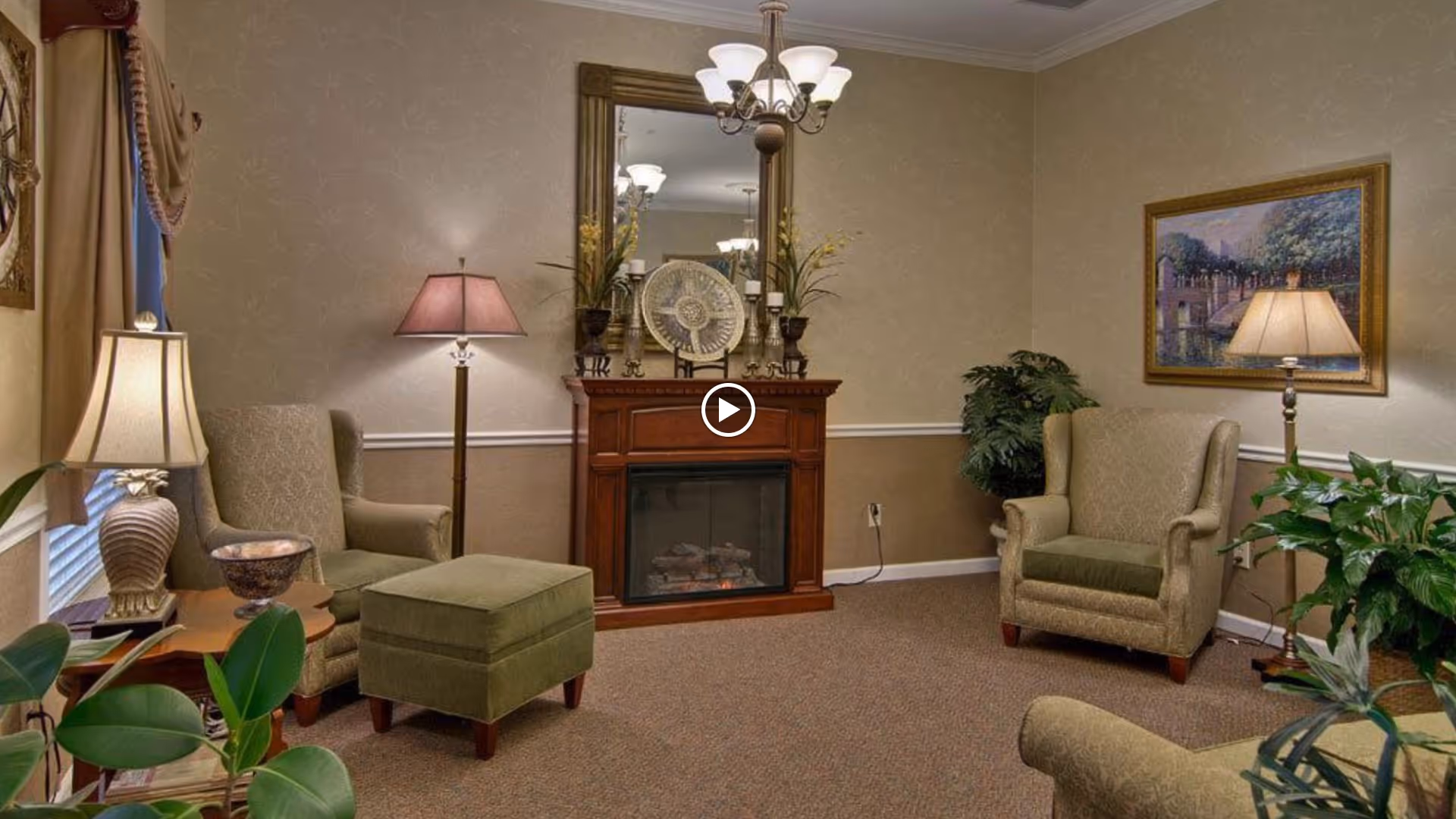 Cozy living room with upholstered armchairs, an ottoman, a fireplace with a mirror, and several lamps.