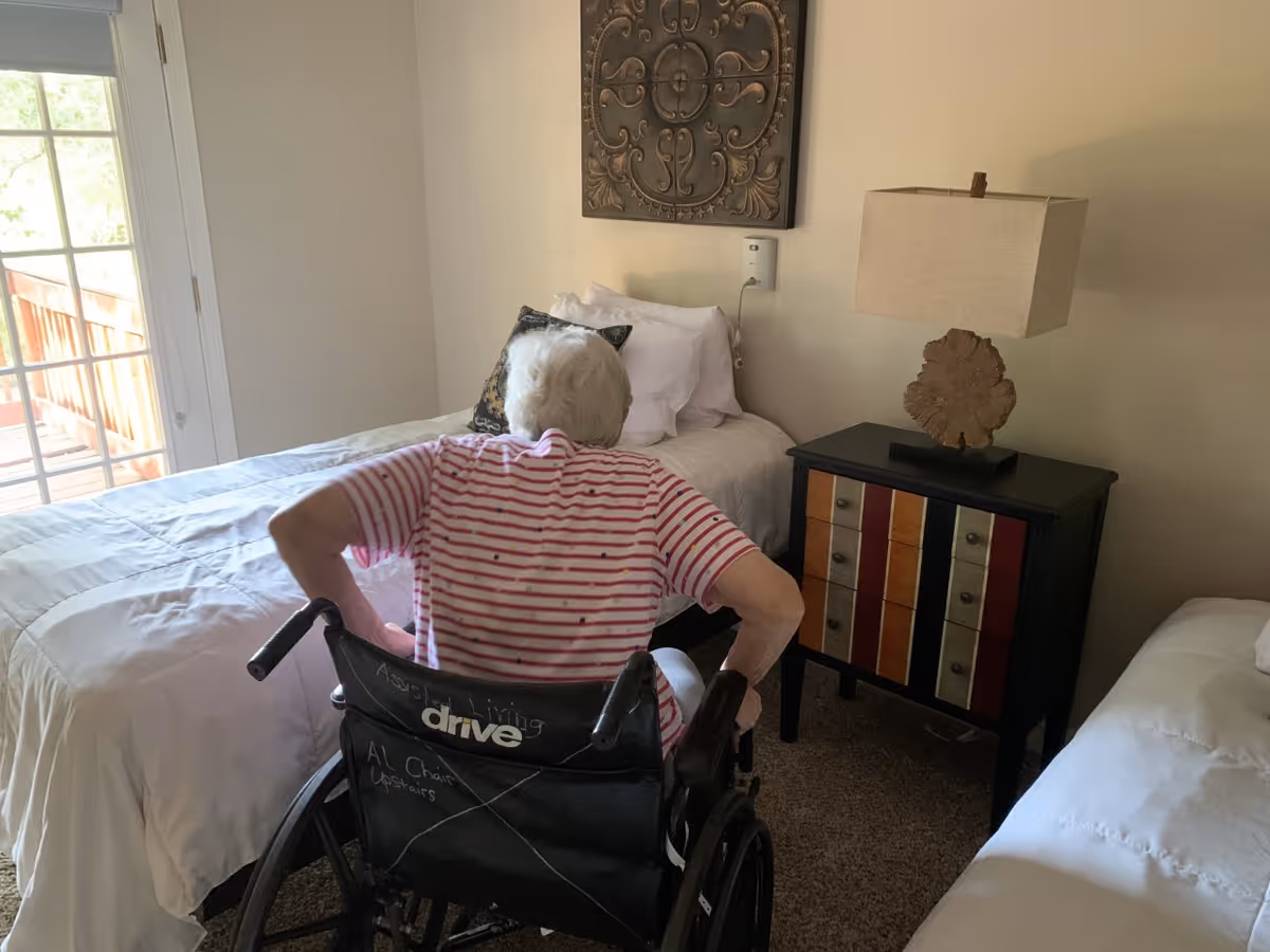 An elderly person in a wheelchair facing a bed in a bedroom with light-colored walls. The room has a bedside table with a lamp and decorative wall art above the bed. There is a glass door leading to an outdoor area.