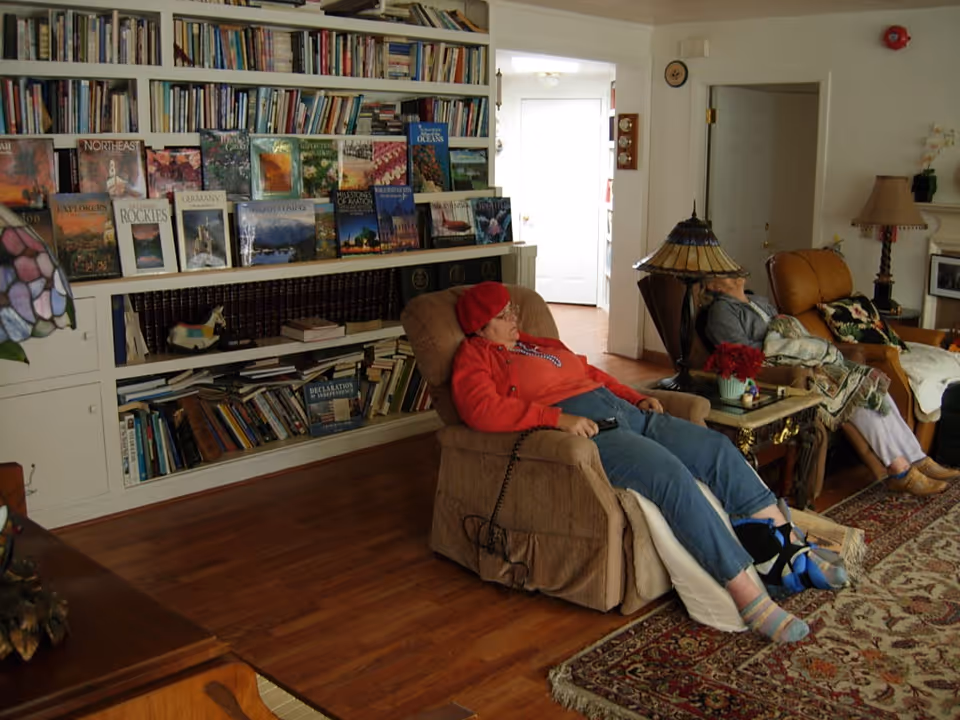 A cozy living room with two elderly women resting in recliner chairs. One woman is wearing a red hat and red sweater, reclining with her feet elevated. The other woman is seated in a brown recliner with a blanket over her legs. Behind them is a large bookshelf filled with books and magazines. The room has wooden flooring, a patterned area rug, a side table with a lamp and flowers, and a doorway leading to another room.