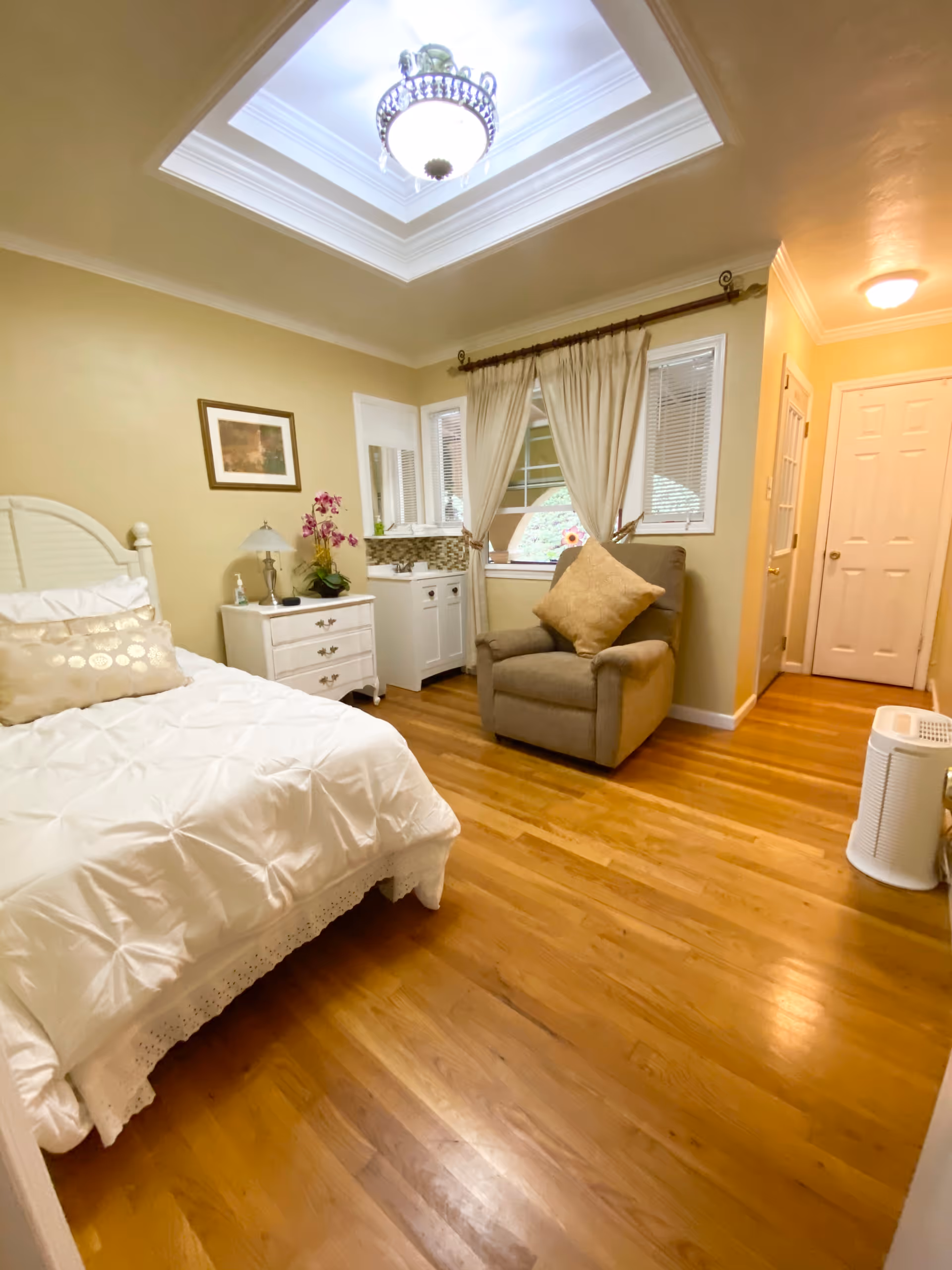 A cozy bedroom with a white bedspread on a bed, a white nightstand with a lamp and a pink orchid, a comfortable armchair with a beige pillow, windows with cream-colored curtains, hardwood floors, and a ceiling light fixture with a skylight above.