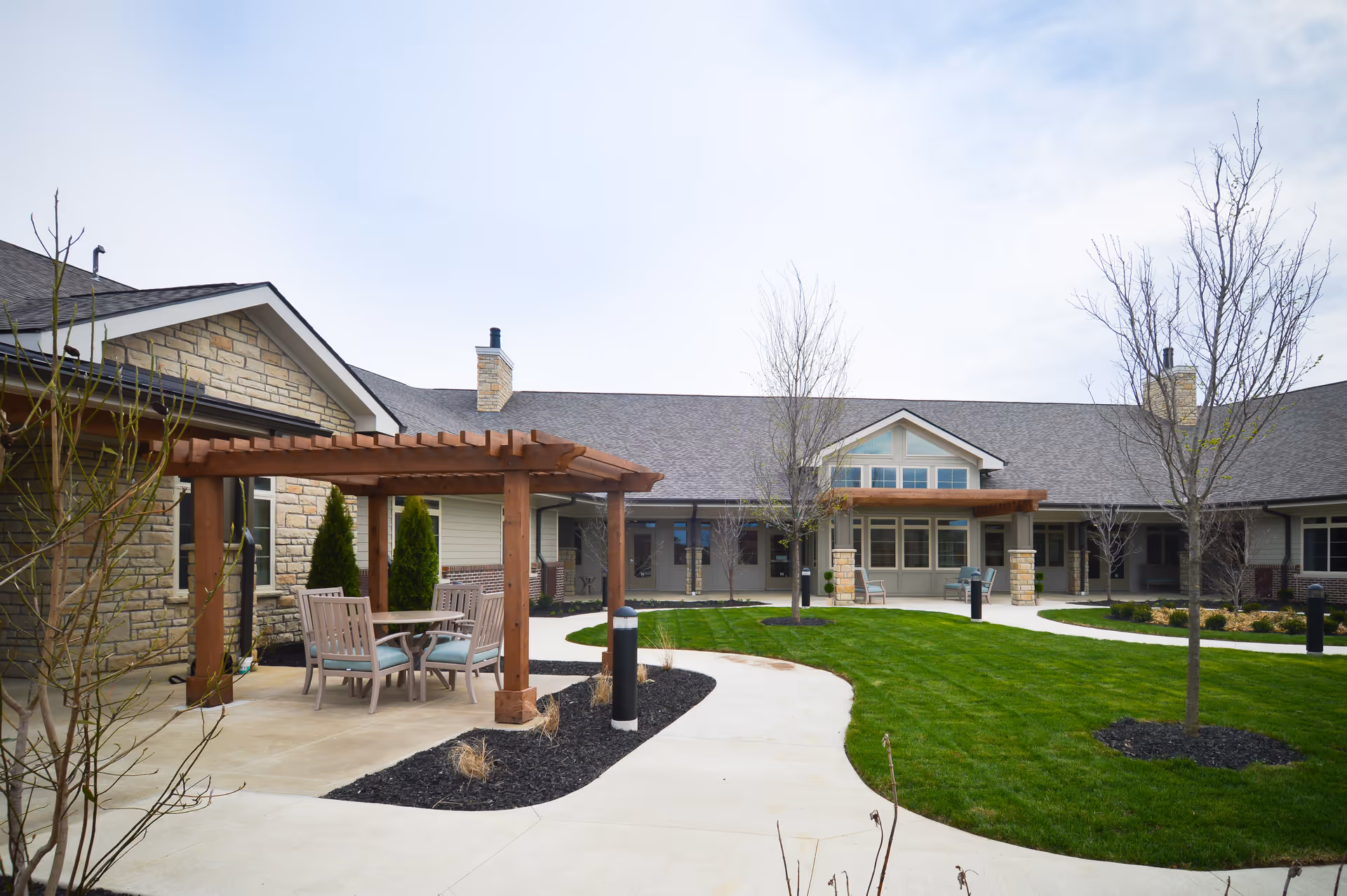 Outdoor courtyard area of The Ganzhorn Suites featuring a wooden pergola with a table and chairs underneath, surrounded by a well-maintained lawn, young trees, and a paved walkway leading to the building entrance.