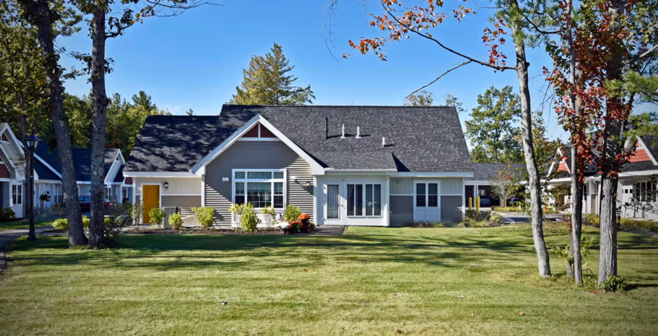 Single-story residential building with gray siding, white trim, and a dark shingled roof, surrounded by a well-maintained lawn and trees under a clear blue sky.