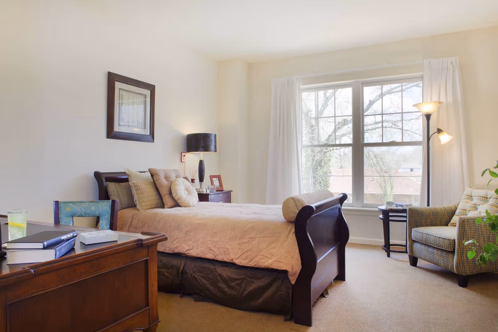 A bright bedroom with a large window covered by white curtains, a wooden bed with beige bedding and multiple pillows, a wooden nightstand with a lamp and picture frame, a patterned armchair with a pillow, a floor lamp, and a wooden desk with books and a glass of water.