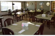 A dining room with multiple tables and chairs arranged neatly. Each table has placemats, napkins, and small centerpieces. Large windows allow natural light to fill the room, and framed pictures and a clock are visible on the walls.